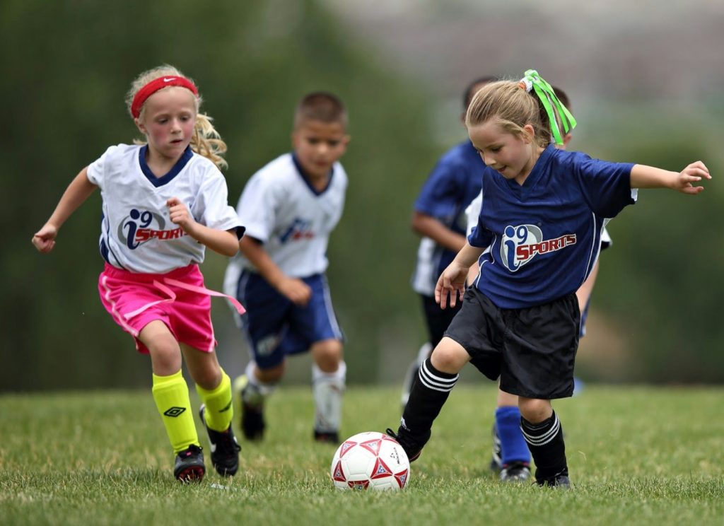 Two young girls in i9 Sports jerseys compete over a soccer ball as their teammates can be seen in the background.