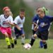 Two young girls in i9 Sports jerseys compete over a soccer ball as their teammates can be seen in the background.