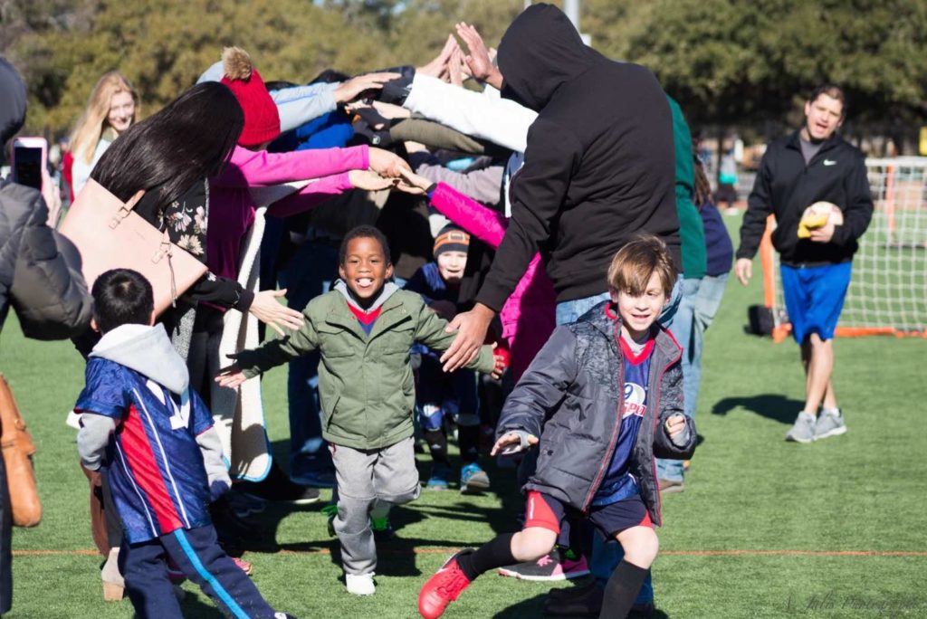 Kids in jackets and i9 Sports soccer uniforms smile and run through a tunnel formed from their parents’ outstretched arms.