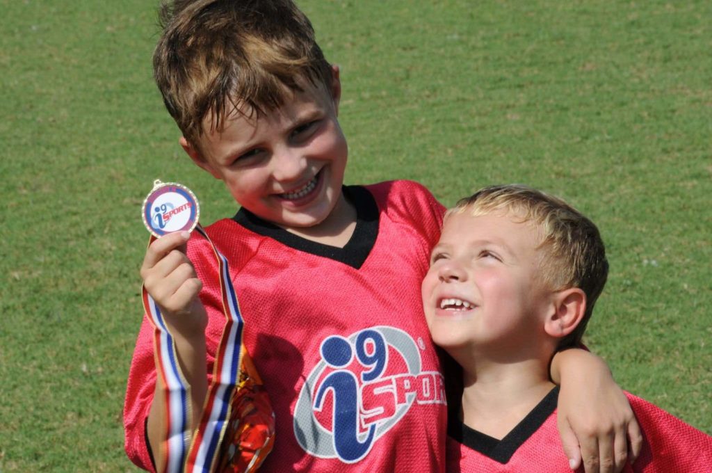 One smiling kid in an i9 Sports uniform is holding a medal and has his arm around another kid, who is smiling at the first kid.