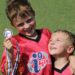 One smiling kid in an i9 Sports uniform is holding a medal and has his arm around another kid, who is smiling at the first kid.