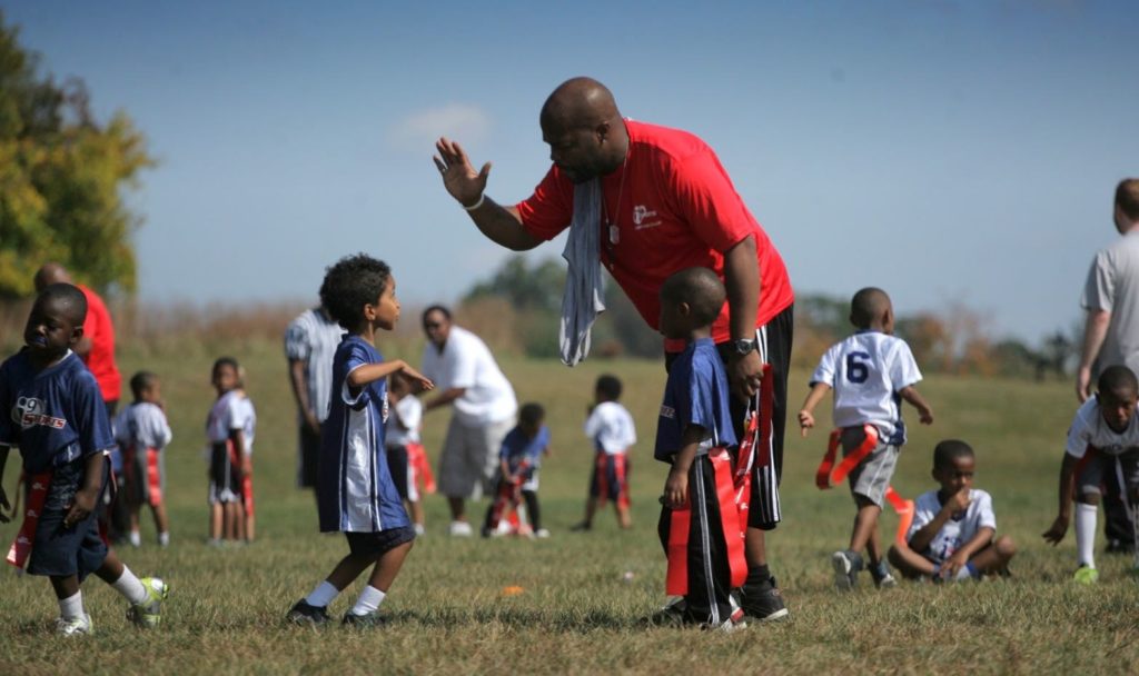 A man in a red i9 Sports t-shirt bends over to give a high five to a kid in an i9 Sports football jersey as other flag football players mill about the field.