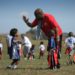 A man in a red i9 Sports t-shirt bends over to give a high five to a kid in an i9 Sports football jersey as other flag football players mill about the field.