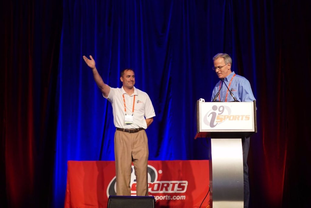 i9 Sports multi-unit franchisee Jeff Mackey stands on stage and waves next to i9 Sports CEO Brian Sanders, who is standing at a podium nearby.