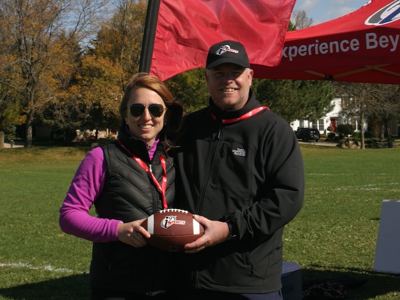 i9 Sports franchise owners Rachel and Michael DuPont hold an i9 Sports-branded football while standing in a grassy sports field by a red flag and tent.