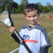 A smiling kid in an i9 Sports jersey stands on a field while holding a lacrosse stick.