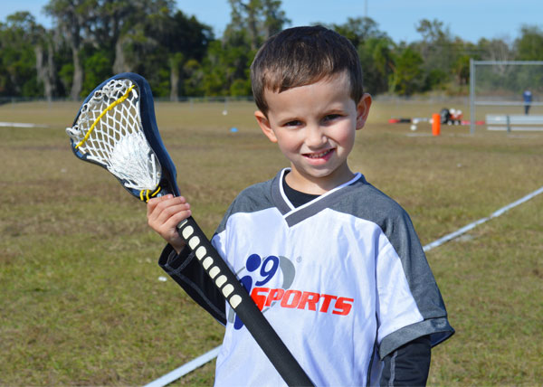 A smiling kid in an i9 Sports jersey stands on a field while holding a lacrosse stick.