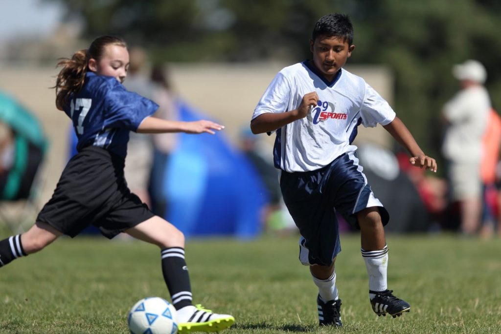 A girl in a blue i9 Sports jersey and a boy in a white i9 Sports jersey compete for a soccer ball on a grassy field.