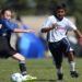 A girl in a blue i9 Sports jersey and a boy in a white i9 Sports jersey compete for a soccer ball on a grassy field.