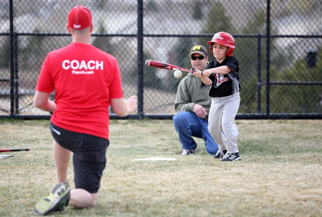 A small boy in an i9 Sports baseball uniform and helmet swings a baseball bat at a pitch. A man is kneeling behind home plate as a catcher, and a pitcher in a red “Coach” t-shirt is on one knee in the foreground.