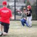 A small boy in an i9 Sports baseball uniform and helmet swings a baseball bat at a pitch. A man is kneeling behind home plate as a catcher, and a pitcher in a red “Coach” t-shirt is on one knee in the foreground.