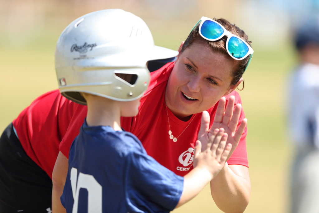 A woman in a red T-shirt high-fives a young boy in a helmet and blue jersey.