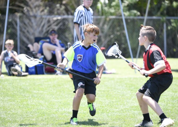 Two boys, one in a blue and green jersey and one in a red and black jersey, spar on a grassy field with lacrosse sticks.