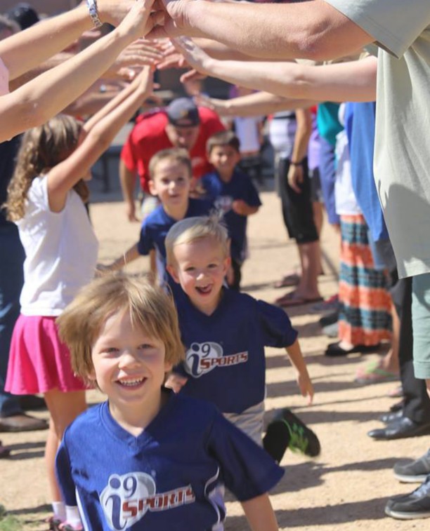 Little children in blue i9 Sports jerseys run beneath a tunnel formed by the upraised arms of two rows of bigger children. 