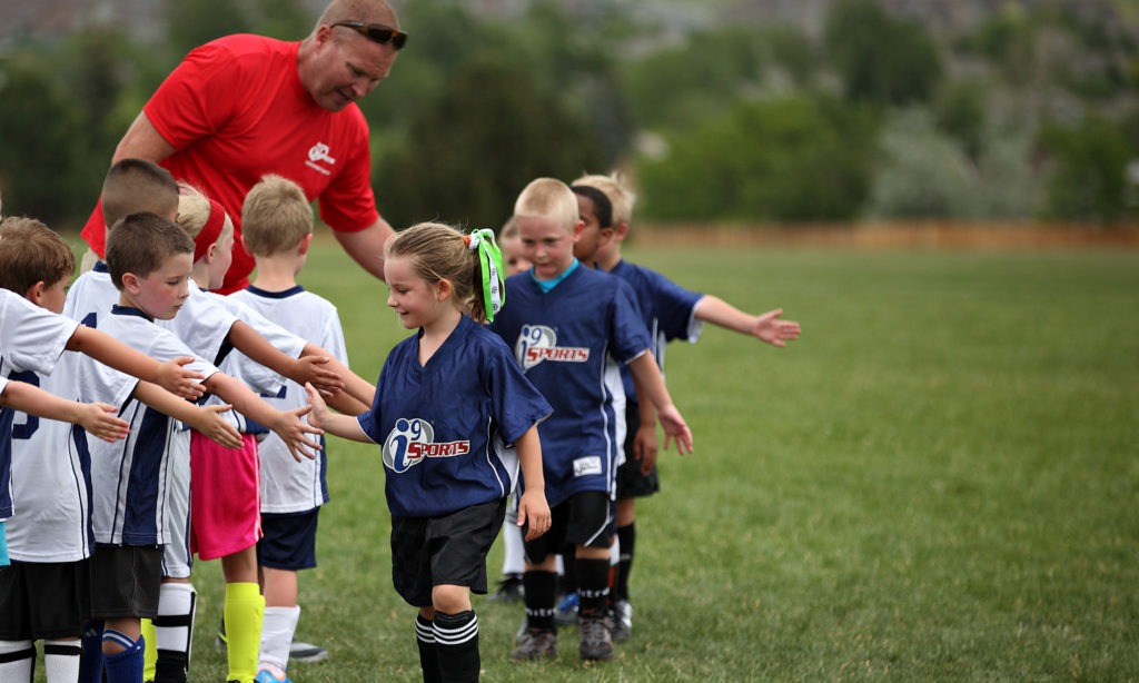 A coach in a red shirt leans over to high five a group of children in blue i9 Sports jerseys walking in a line in a grassy field.
