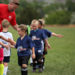 A coach in a red shirt leans over to high five a group of children in blue i9 Sports jerseys walking in a line in a grassy field.