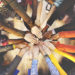 A photo from above shows a variety of people standing in a circle with their arms stretched the center and their hands overlapping or touching.