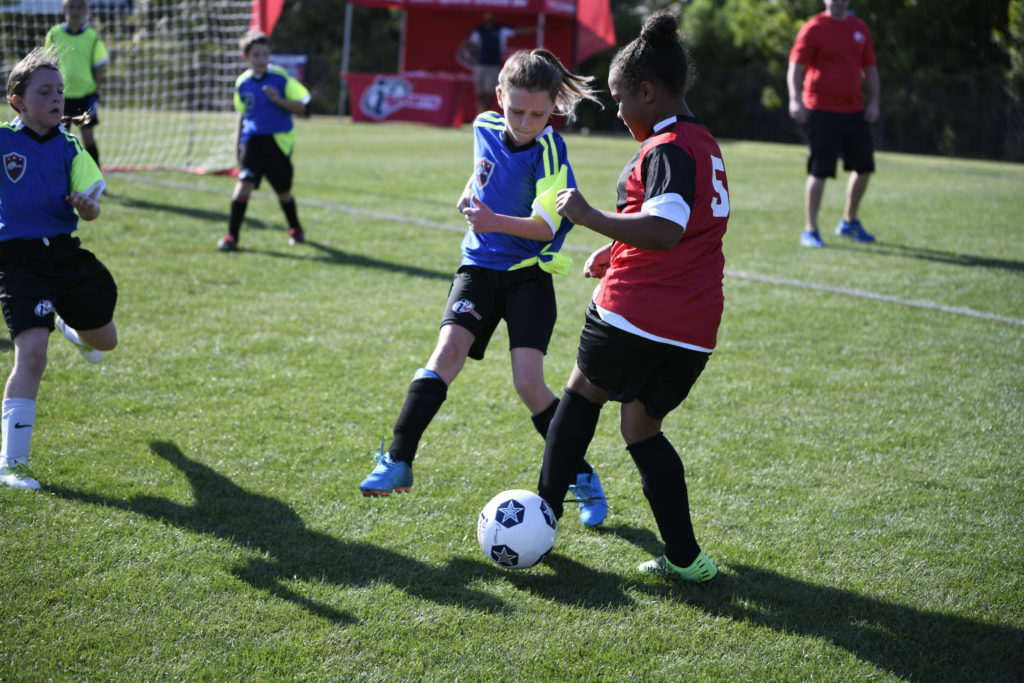 Girls from opposing soccer teams, wearing a red or blue i9 Sports jersey, kick at a soccer ball on a grassy field while other kids run around them.