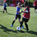 Girls from opposing soccer teams, wearing a red or blue i9 Sports jersey, kick at a soccer ball on a grassy field while other kids run around them.