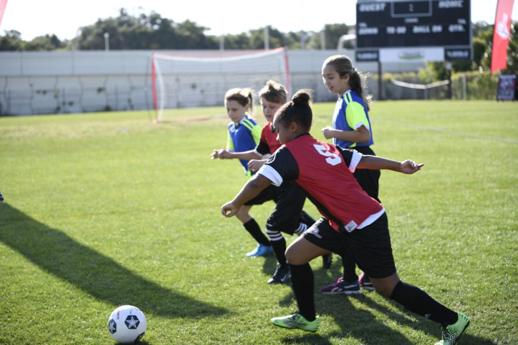Boys and girls in blue and red jerseys rush toward a soccer ball on a grassy green field.