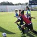 Boys and girls in blue and red jerseys rush toward a soccer ball on a grassy green field.