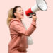 A ponytailed woman in a dusty pink hoodie appears to be yelling through a megaphone.