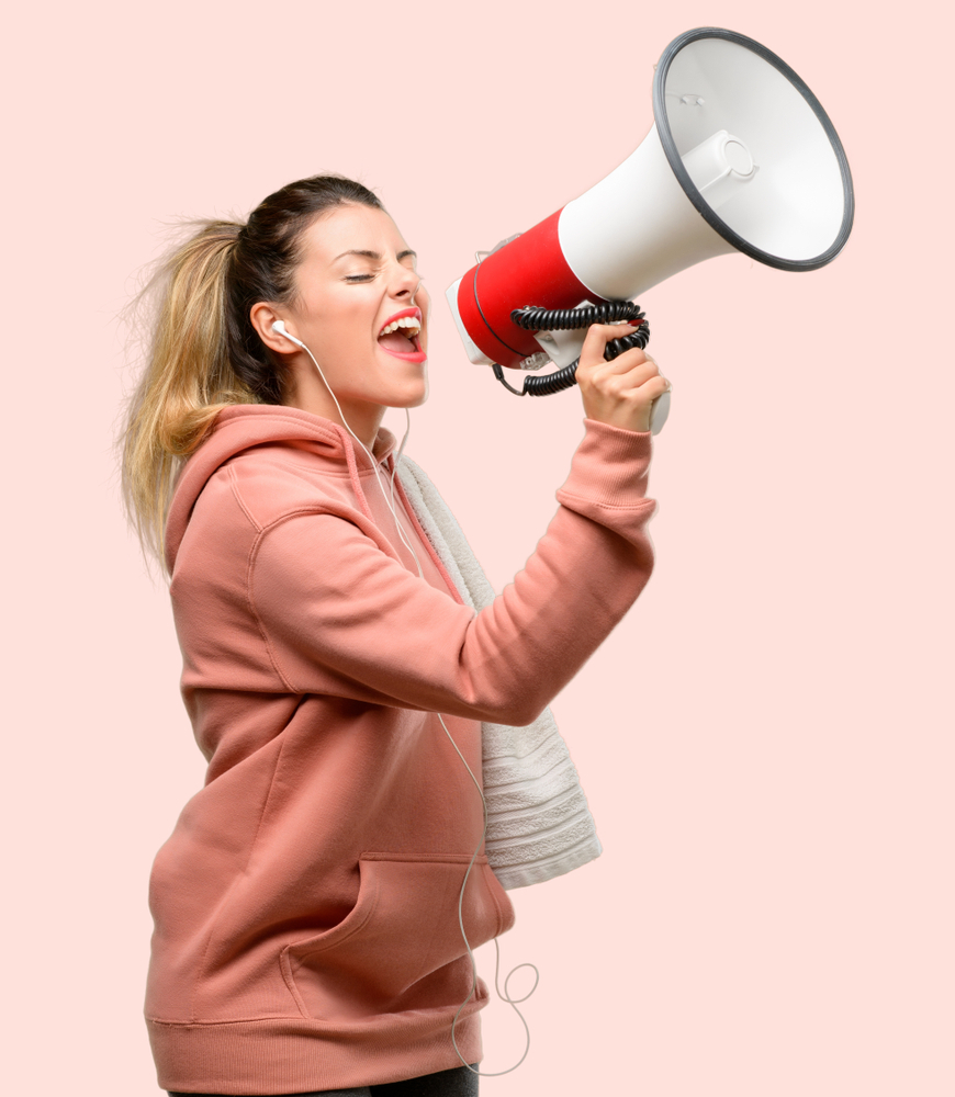 A ponytailed woman in a dusty pink hoodie appears to be yelling through a megaphone.