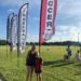 A blonde mom in sunglasses has an arm around her young daughter standing on the soccer field under a banner that says "Soccer." In a row behind that banner are banners that read "Baseball" and "Lacrosse" and a third banner whose sport is hidden. All have the i9 Sports franchise logo on them.