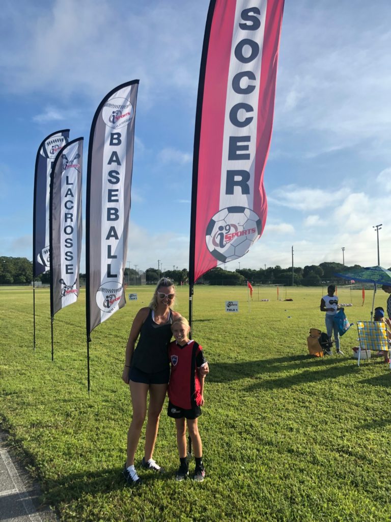 A blonde mom in sunglasses has an arm around her young daughter standing on the soccer field under a banner that says "Soccer." In a row behind that banner are banners that read "Baseball" and "Lacrosse" and a third banner whose sport is hidden. All have the i9 Sports franchise logo on them.