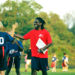 A coach in a red shirt high fives a flag football youth player in a blue jersey, standing on a field among other flag football players facing away from the camera.