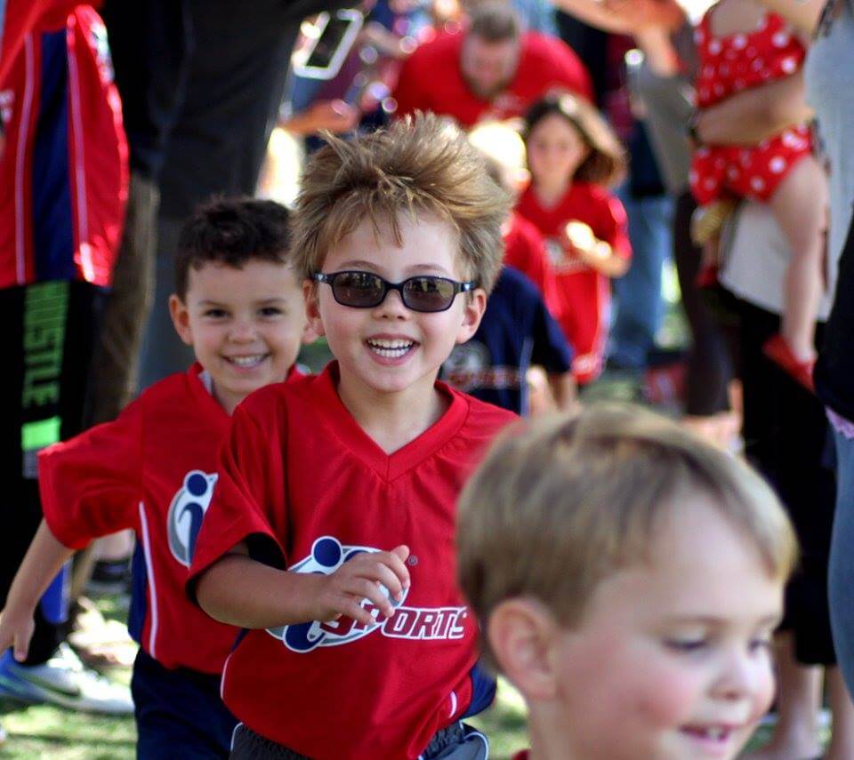 ALT: Three little boys in i9 Sports jerseys run onto a field, smiling toward the camera, with the boy in the middle wearing Wayfarer-style sunglasses.