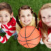 A boy and two girls standing on a swath of green turf hold a soccer ball, basketball and volleyball as they smile up at the camera.