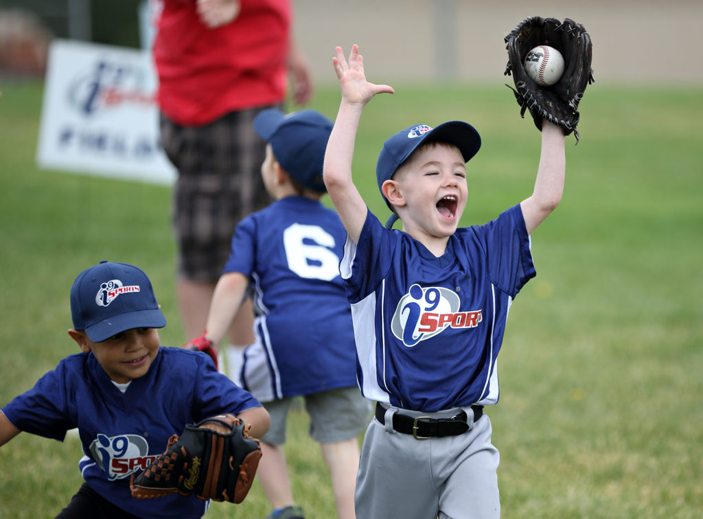 A boy wearing an i9 Sports jersey catches a baseball in his mitt with a look of joy on his face while a teammate looks on.