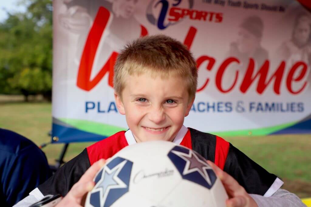 A boy holding a soccer ball in front of him smiles at the camera.