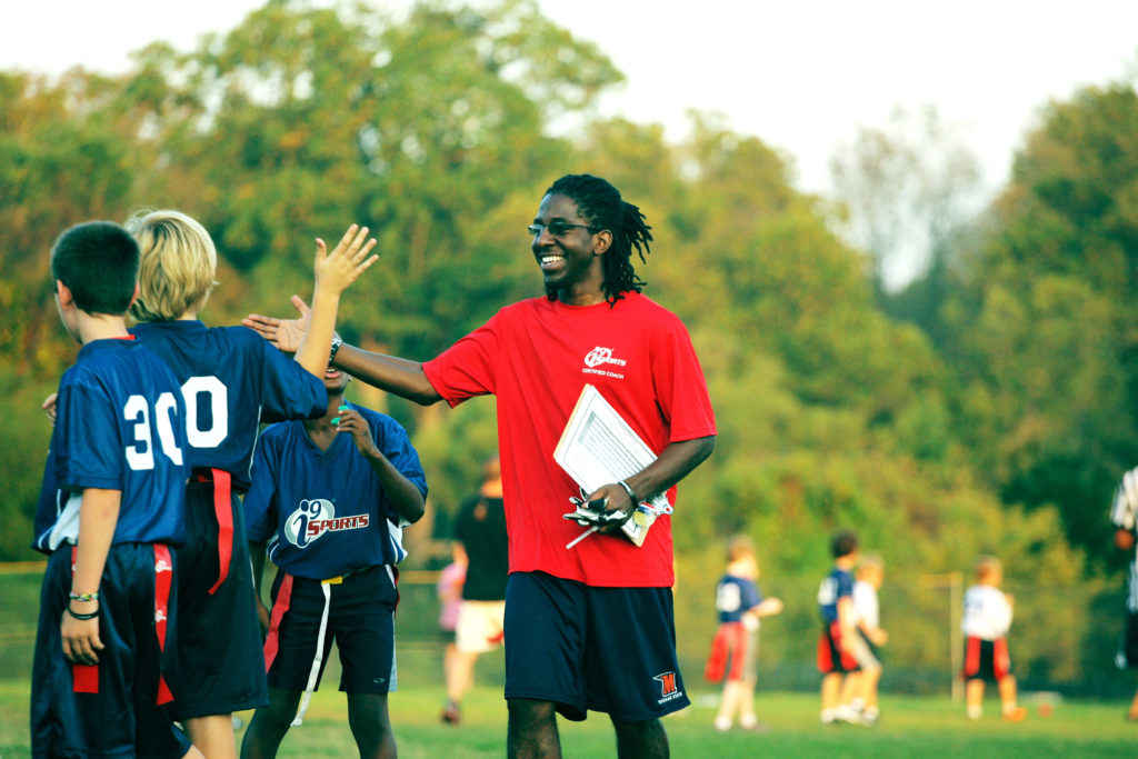 A smiling coach in a red i9 Sports jersey carrying a clipboard gives a high five to kids on a flag football field