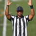 A referee in a black-and-white striped shirt holds up both arms in the symbol for a touchdown.