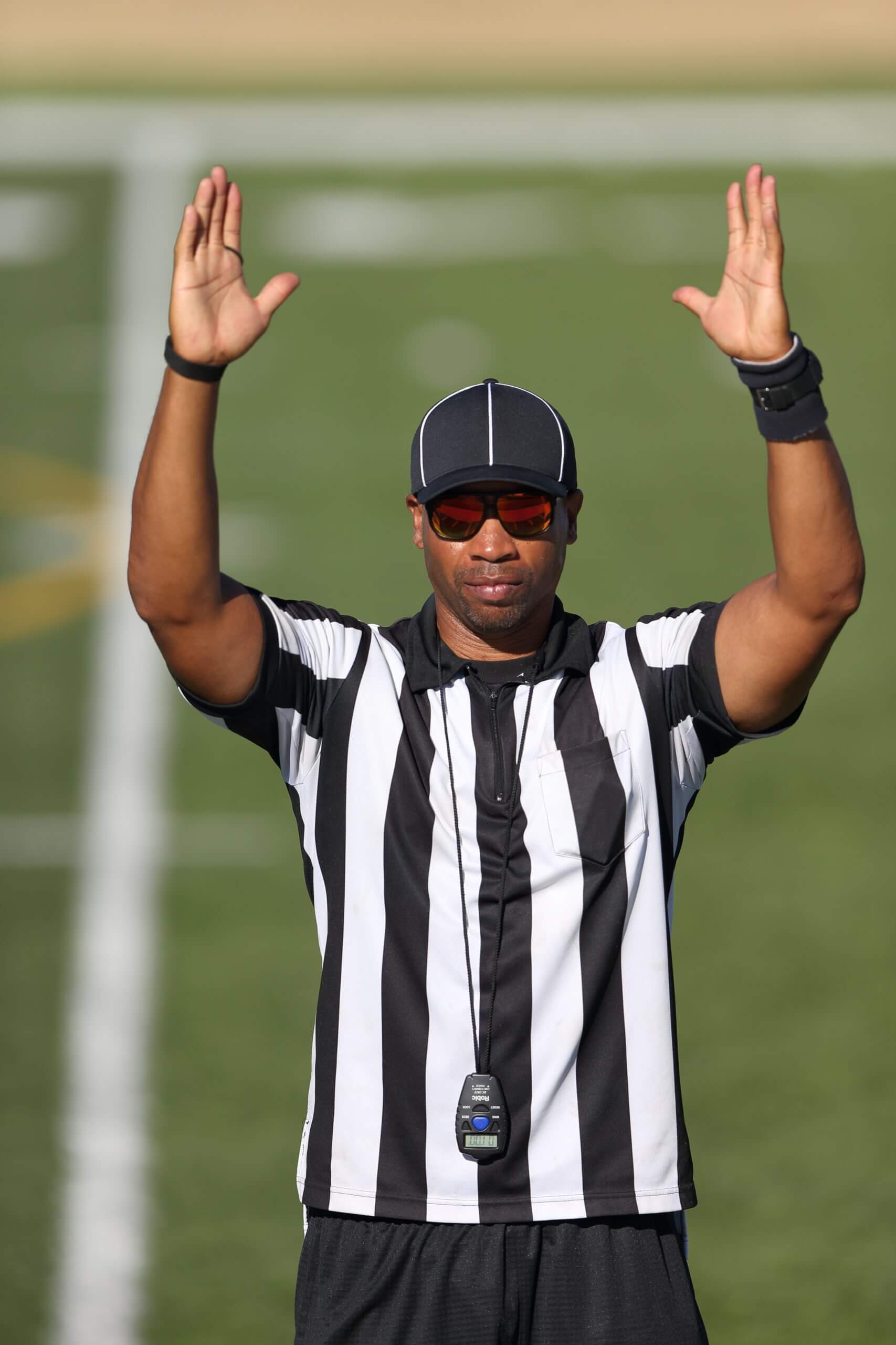 A referee in a black-and-white striped shirt holds up both arms in the symbol for a touchdown.