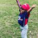 A little boy in a red, white and blue i9 Sports uniform with a baseball cap has both arms outstretched above his head in celebration on a grassy field.