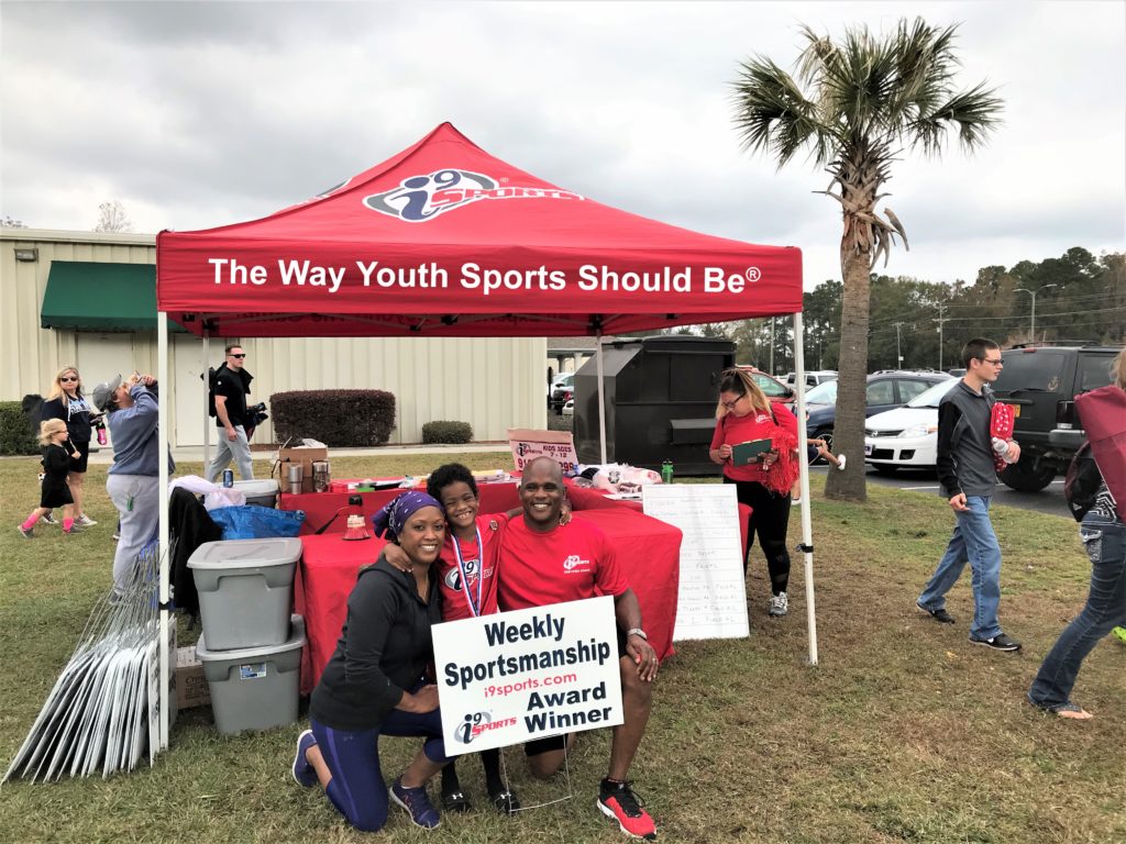 Walter and Sandra Reaves and their young son pose in front of an i9 Sports tent whose awning reads, "The Way Youth Sports Should Be"