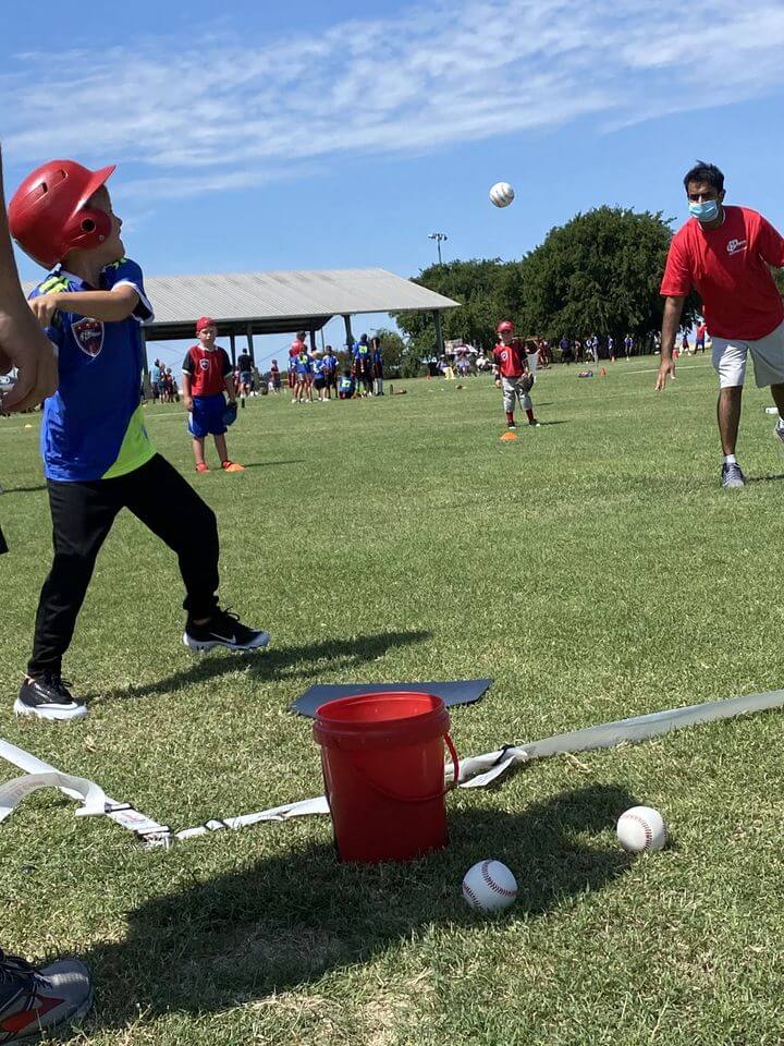 A coach in a mask pitched to a boy in an i9 Sports jersey with a bat.