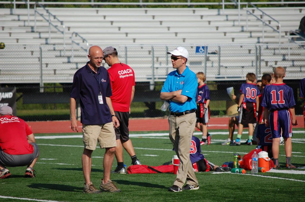 Men in khakis and blue i9 Sports polo shirts talk to each other in the foreground. In the background are children in flag football uniforms and assorted sports equipment on a football field.