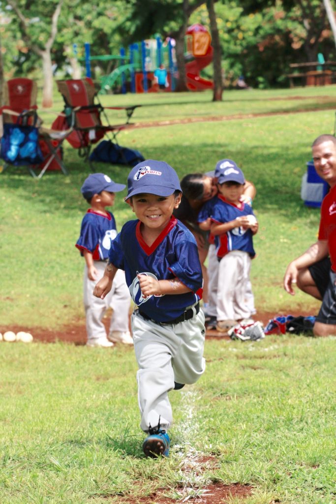 A little boy in an i9 Sports jersey runs down a baseline toward the camera and smiling. In the background, other little boys in i9 Sports uniforms are gathered next to a kneeling coach.