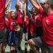 A group of kids in i9 Sports uniforms high-five in a circle with their kneeling coach.