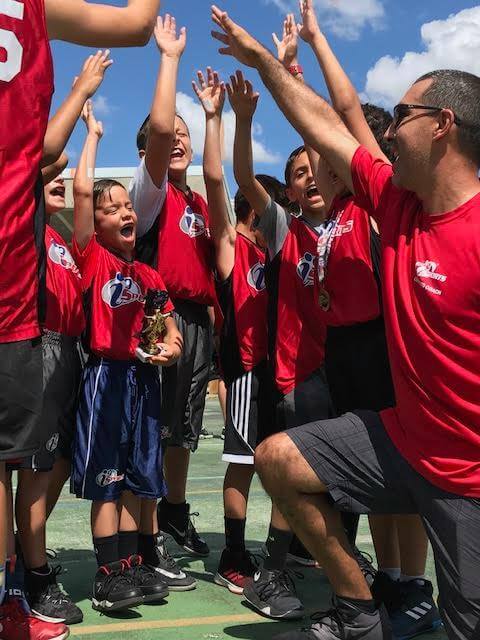 A group of kids in i9 Sports uniforms high-five in a circle with their kneeling coach.