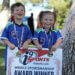 A boy and girl standing beneath a tree and dressed in i9 Sports jerseys hold a sign that reads, “i9 Sports|i9sports.com|Weekly Sportsmanship Award Winner.” Both are wearing medals around their necks and smiling.