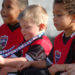 Three little boys in i9 Sports jerseys, one holding a medal around his neck to admire, are sitting together on a bench.