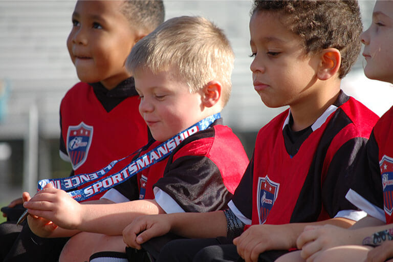 Three little boys in i9 Sports jerseys, one holding a medal around his neck to admire, are sitting together on a bench.