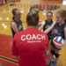 Four young girls gather around a volleyball coach inside a gym.