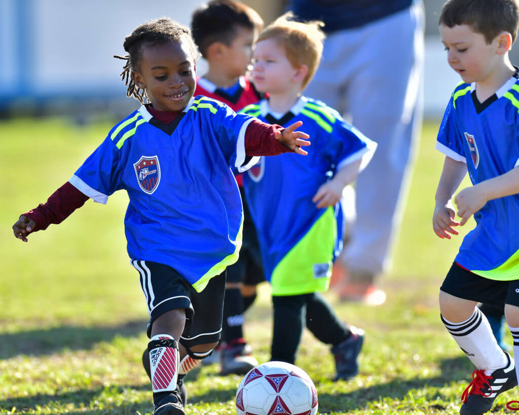 Four young children play soccer with a coach in the background. A young African-American girl is smiling and about to kick the soccer ball.