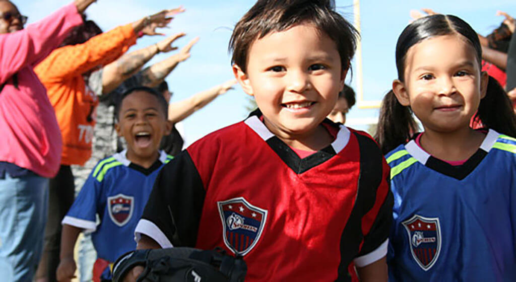 Three kids - a boy and a girl with another boy behind them - run through a parent tunnel, which is a group of parents standing on either side of the kids with their arms raised.